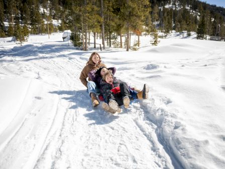 Two people ride a sled down a snowy slope, smiling, as forested hills and fresh powder surround them.