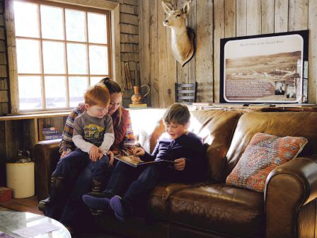 Three kids read together on a brown leather couch in a sunlit cabin, with a deer mount and framed display on the wall nearby.