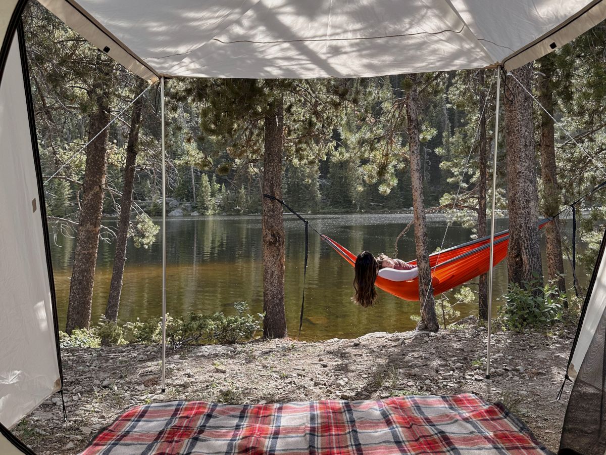 A camping scene: view from a tent entrance shows a plaid blanket inside, trees and a lake beyond, with a red hammock tied between them.