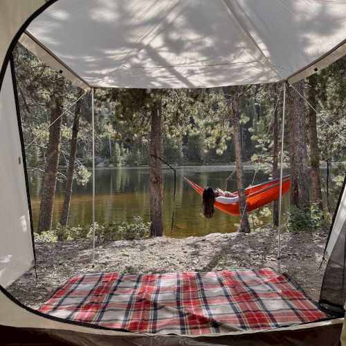 Looking out from a tent at a lakeside campsite; a red hammock hangs between trees, plaid blanket inside the tent, peaceful nature vibe.