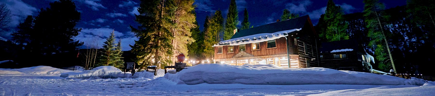A cozy wooden cabin glows warmly in a snowy night, tall pines nearby, starry sky above, and freshly plowed paths curving through the snow.