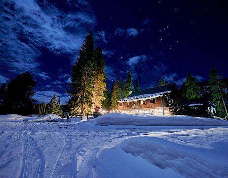 A cozy wooden cabin glows warmly in a snowy night, tall pines nearby, starry sky above, and freshly plowed paths curving through the snow.