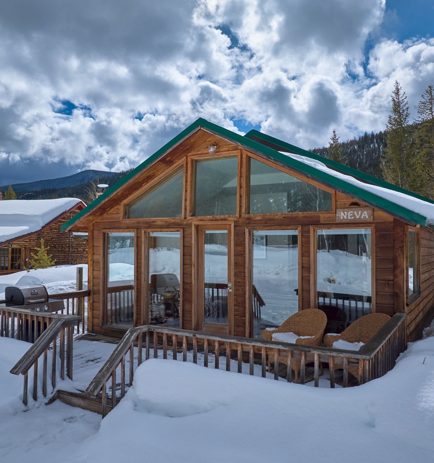 A cozy log cabin with large glass front, snow around, wooden deck and chairs, set in a wintery mountain scene.
