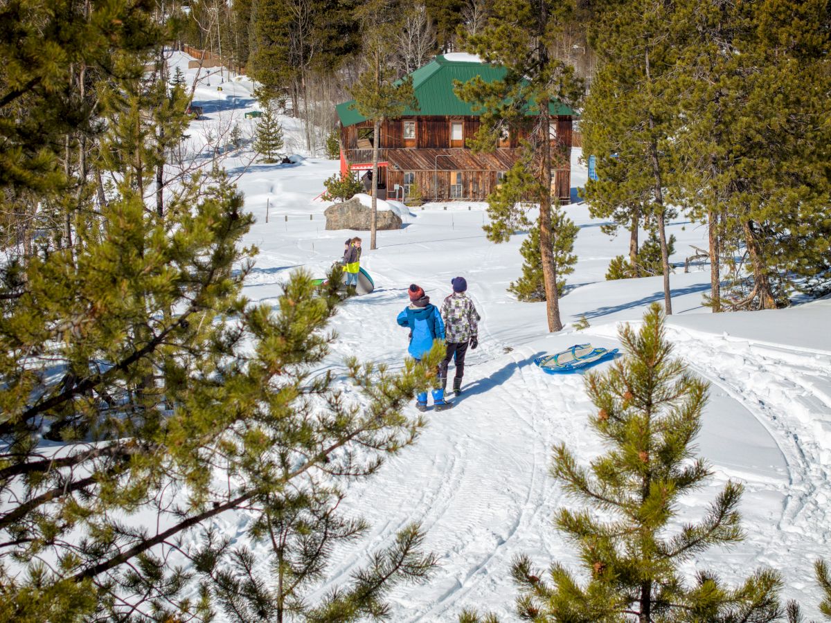 A snowy mountain scene with three people skiing near a wooden cabin, pine trees lining a groomed trail, and a green roof building in the background.