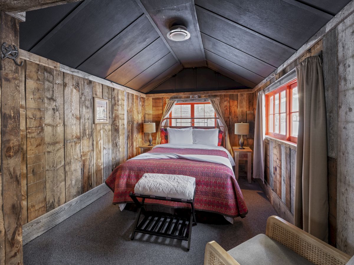 A cozy rustic bedroom in an attic with wooden panel walls, a red bedspread, two bedside lamps, and a small bench at the foot of the bed.