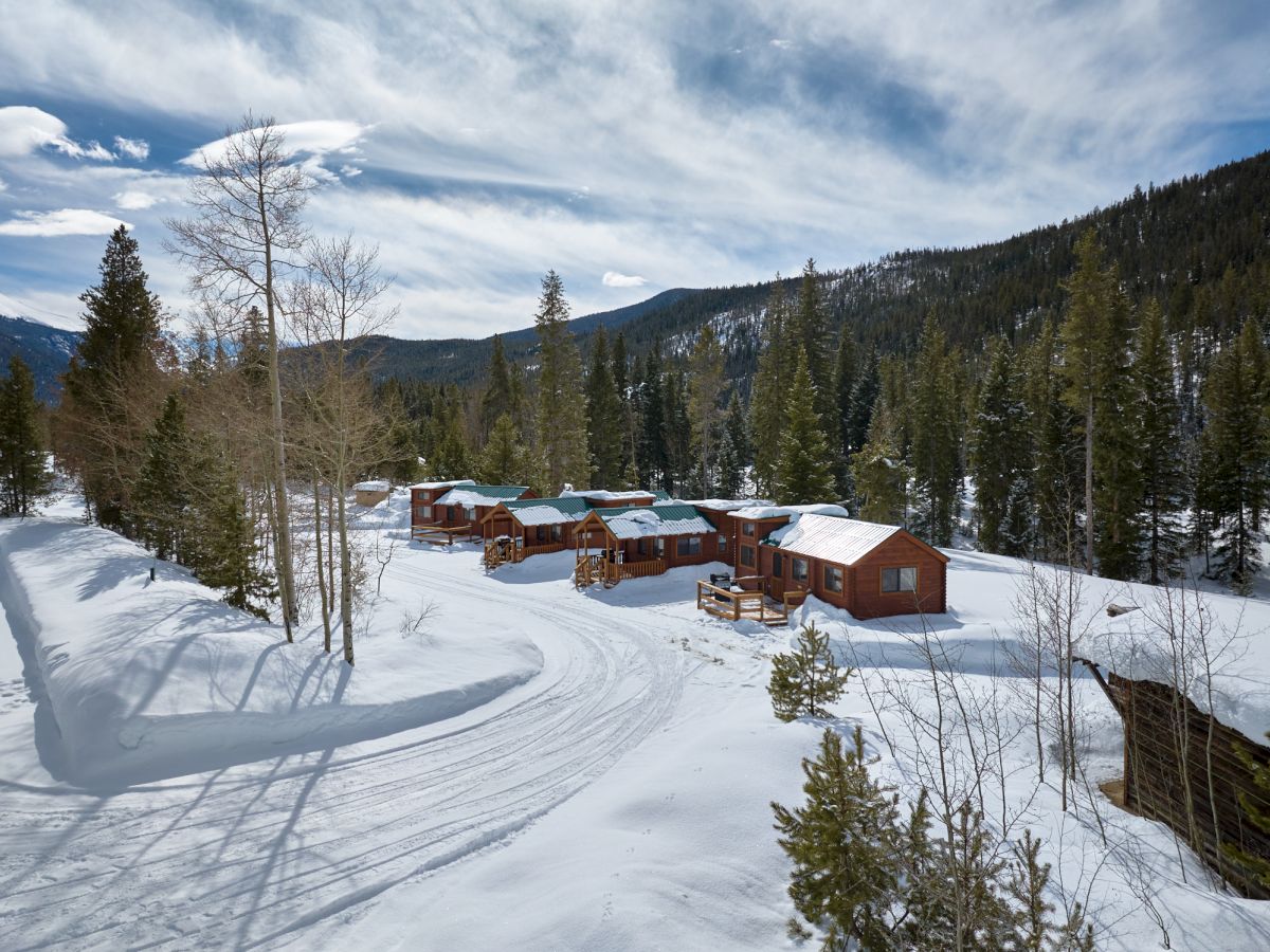 Snowy mountain cabin village nestled among tall pines, with winding tracks in fresh powder and clear blue sky above.