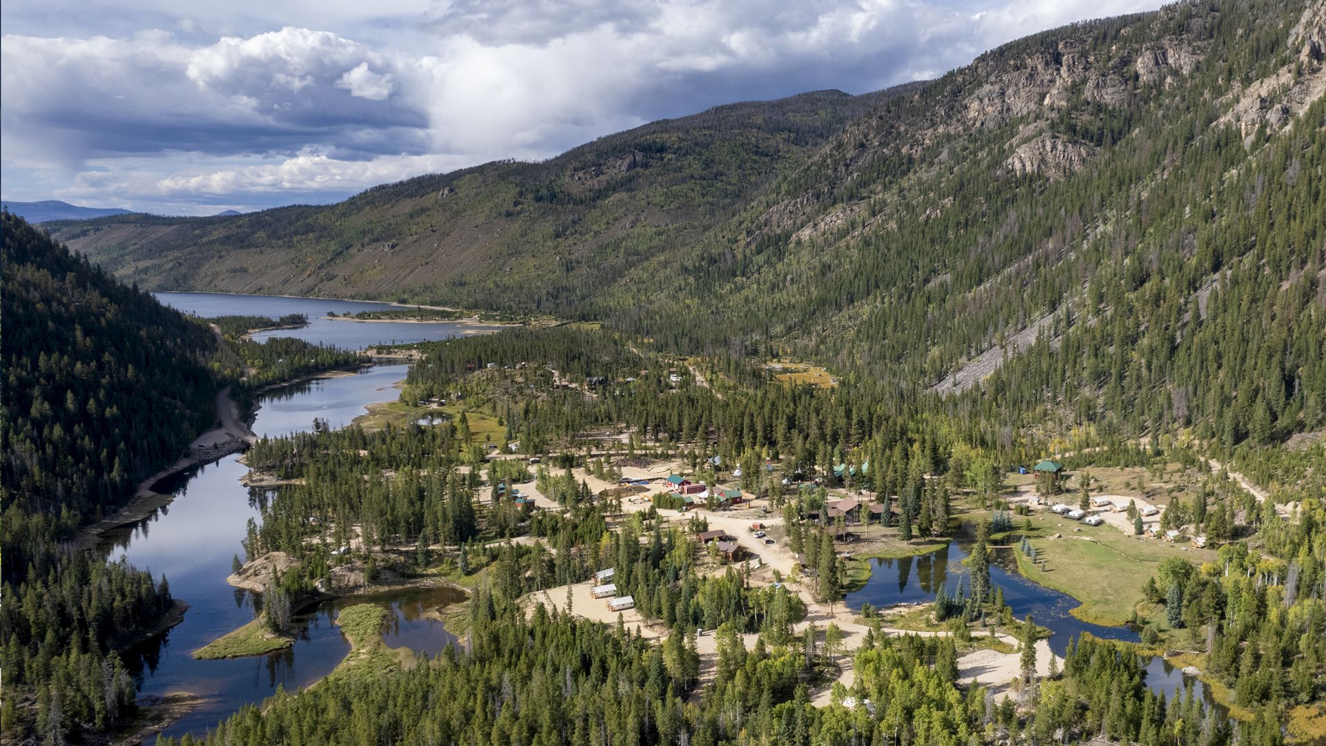 A mountain valley with a winding river, dense evergreen forests, and a small lakeside village nestled among trees, under a partly cloudy sky.