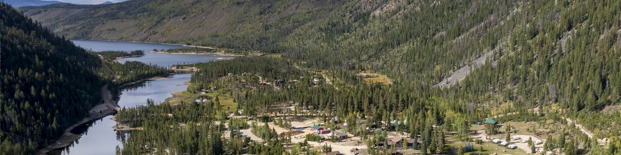 A mountain valley with a winding river, dense evergreen forests, and a small lakeside village nestled among trees, under a partly cloudy sky.