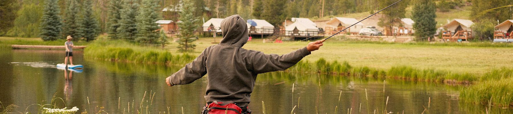 Two people fishing in a lake and a person with arms raised, a grassy bank, trees, and cabins in the background, sunny and tranquil.