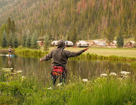 Two people fishing in a lake and a person with arms raised, a grassy bank, trees, and cabins in the background, sunny and tranquil.