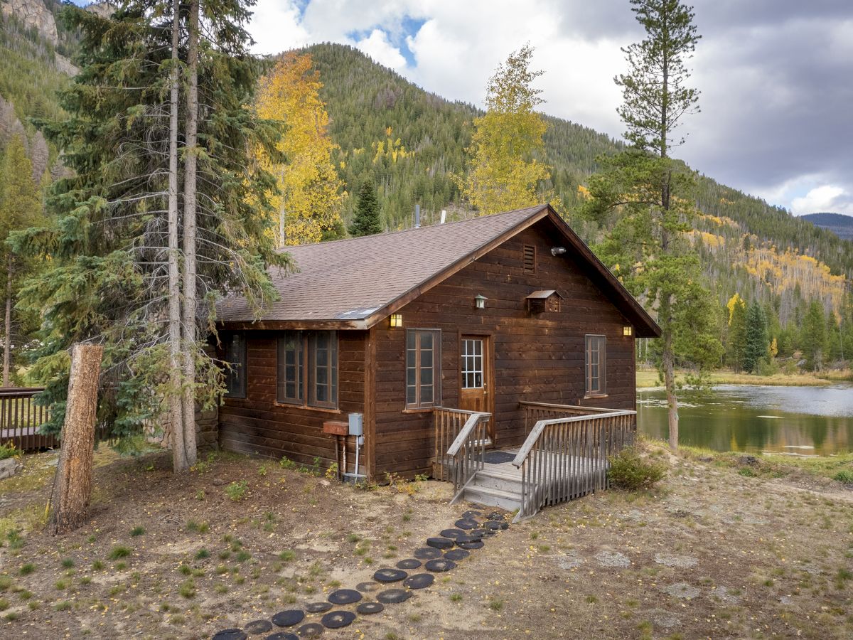 A rustic wood cabin by a lake, nestled among trees and mountains with a small porch and stepping stones leading to the door.