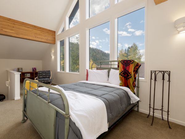 A cozy bedroom with a metal-framed bed, large windows, a wooden beam, and a small seating area in the corner, bright and airy.
