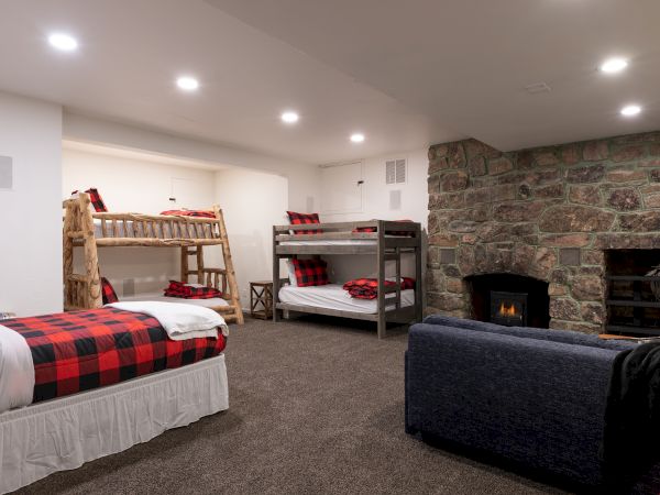 A cozy basement with two bunk beds, a sofa, a stone fireplace, and a small heater area, all under bright ceiling lights.