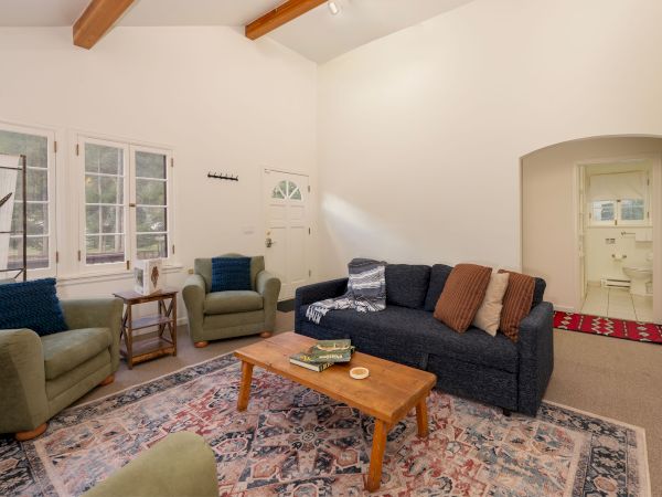 Cozy living room with a dark blue sofa, green armchairs, wooden coffee table, patterned rug, white walls, and exposed ceiling beams. End with a sentence.