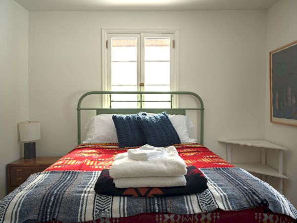 A neatly made bedroom with a green metal headboard, a patterned quilt, two white pillows, stacked towels, a small nightstand with a lamp, and a window at the headboard.