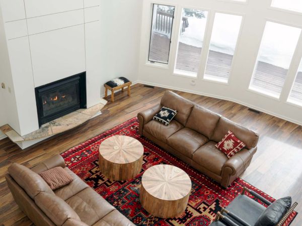A modern living room with a tall white fireplace, large windows, and a red patterned rug. It features three brown leather sofas, two round wooden coffee tables, and a blue armchair set by the couches. The space is airy and bright.