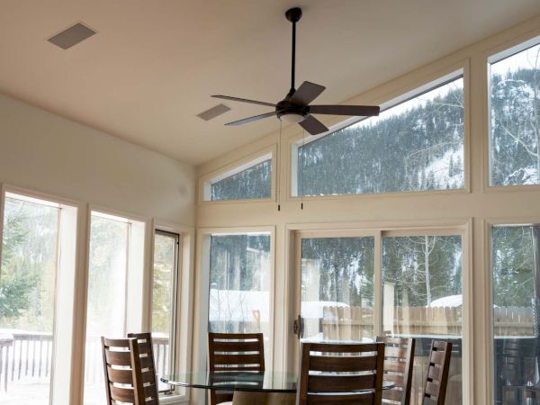 A bright sunlit dining area with a round table and six wooden chairs, surrounded by large windows and a ceiling fan above.