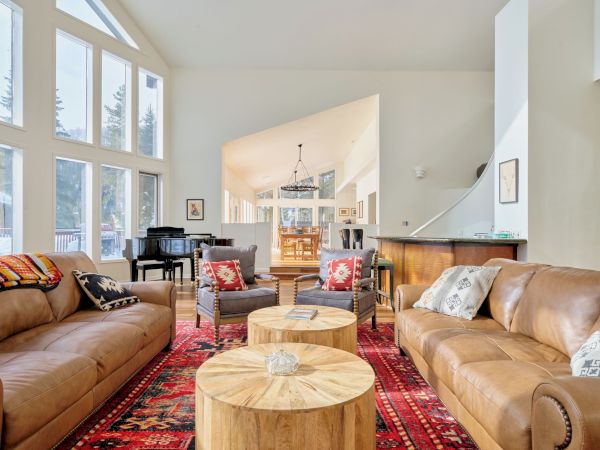 A cozy, mid-century living room with leather sofas, wooden coffee tables, a red patterned rug, piano, and large windows letting in natural light.