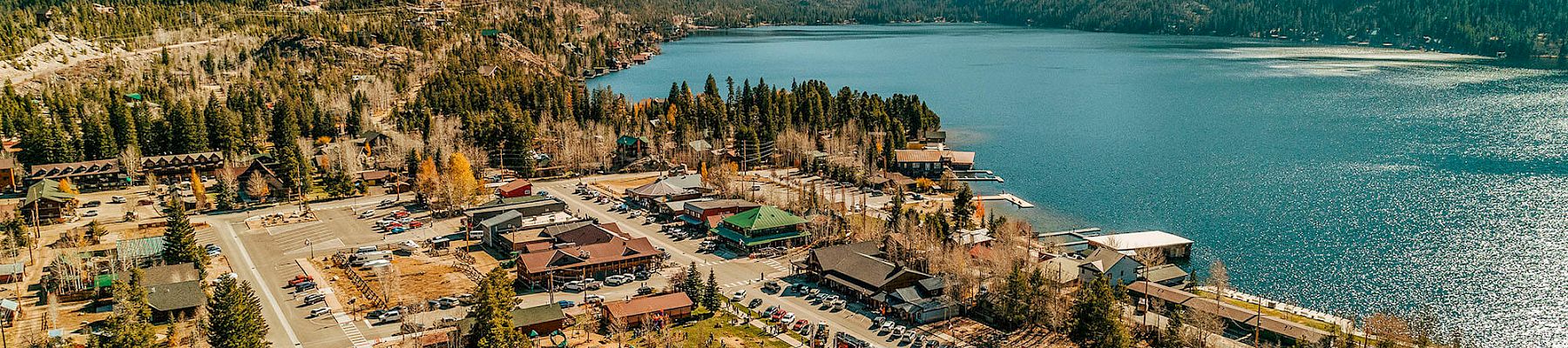 A small lakeside town with a marina, colorful buildings, and a parking lot, nestled among green hills and a calm blue lake.