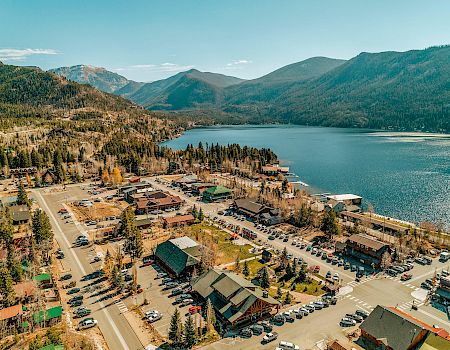 A small lakeside town with a marina, colorful buildings, and a parking lot, nestled among green hills and a calm blue lake.