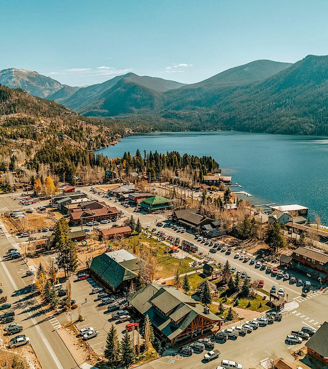 A small lakeside town with a marina, colorful buildings, and a parking lot, nestled among green hills and a calm blue lake.