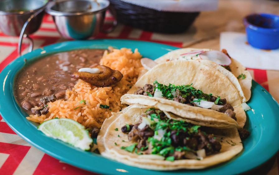 A plate with beef tacos topped with cilantro, lime wedges, and warm tortillas, plus Mexican rice and refried beans on the side. Ending with a period.