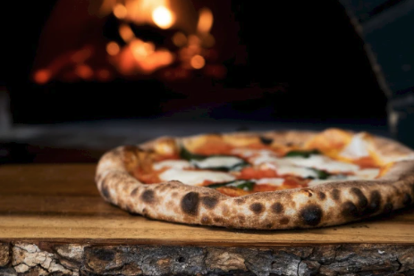 A wood-fired pizza with vegetables sits on a cutting board in front of a glowing oven fire.
