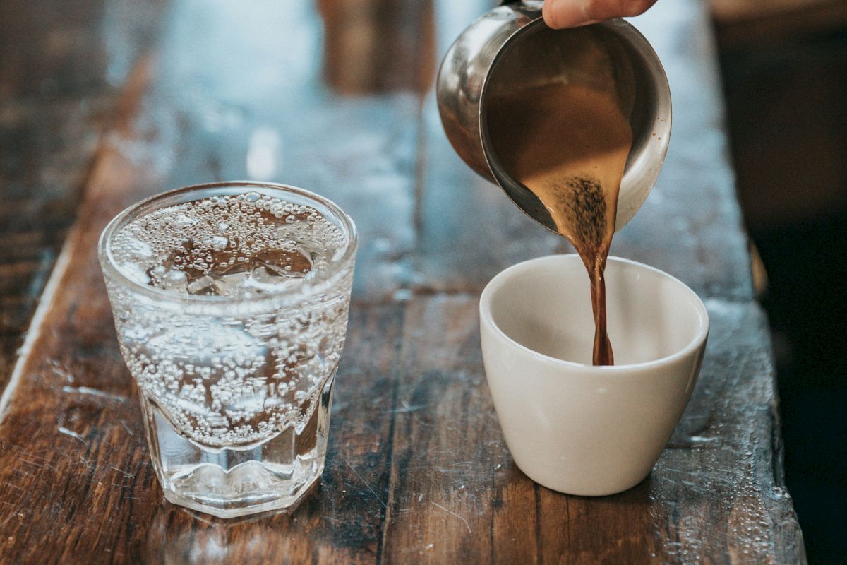 A barista pour-over coffee into a white cup, with a glass of ice water nearby on a wooden counter.