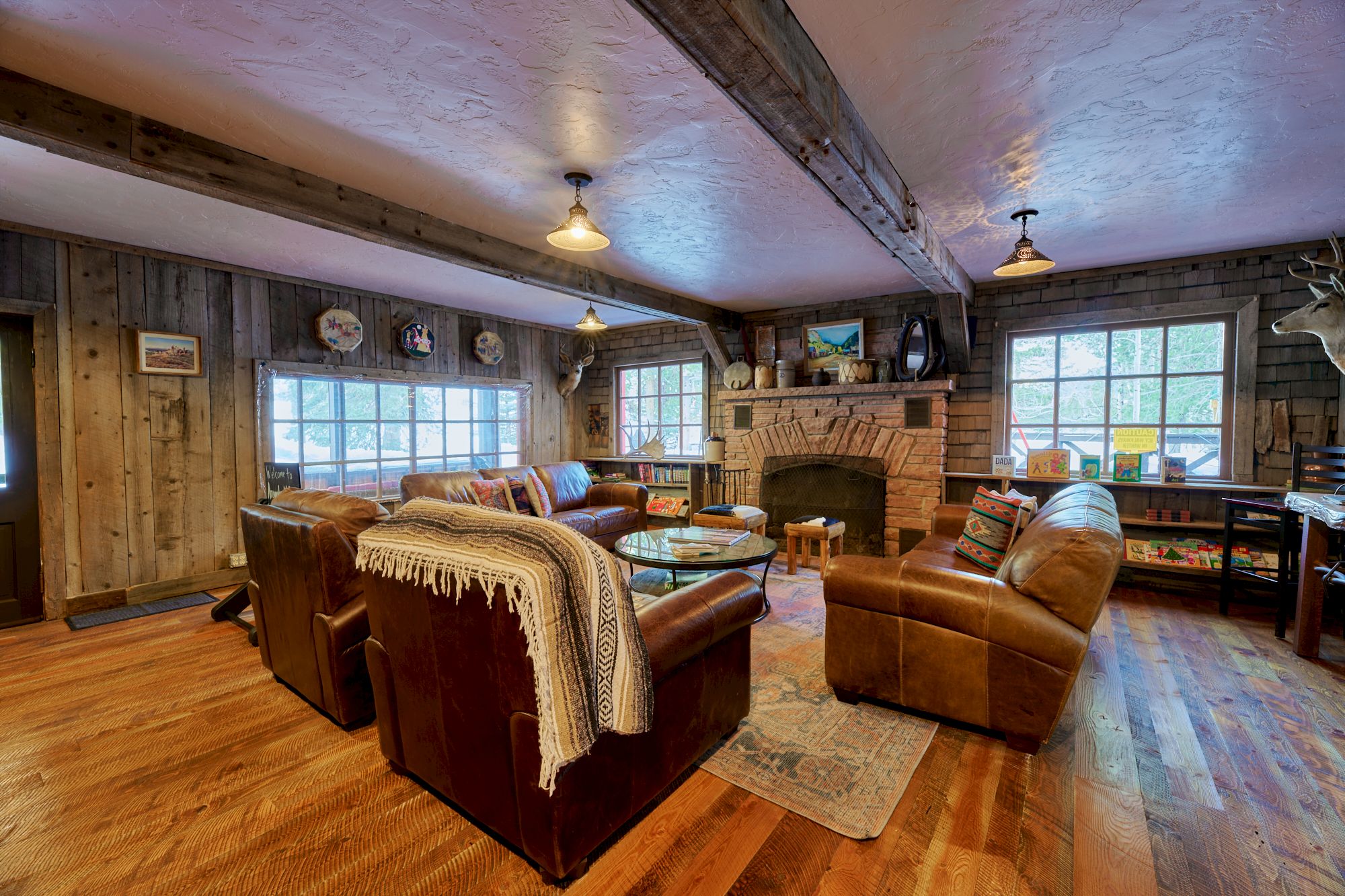 A cozy rustic living room with wooden walls, a stone fireplace, leather sofas, a shaggy throw, warm lighting, and large windows letting in natural light.