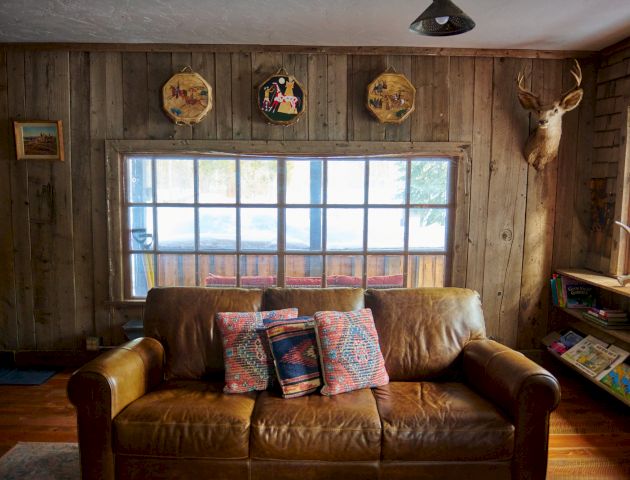 A cozy living room with a window seat, two brown sofas facing a coffee table, warm lighting, and a patterned rug.