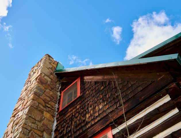 A rustic building with a stone chimney and wooden beams against a bright blue sky. Top it at 140 characters, always ending the sentence.