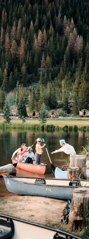 People haul a rowboat ashore by a dock on a calm lake, while others stand and chat near the water&rsquo;s edge, with pine trees and cabins in the background.