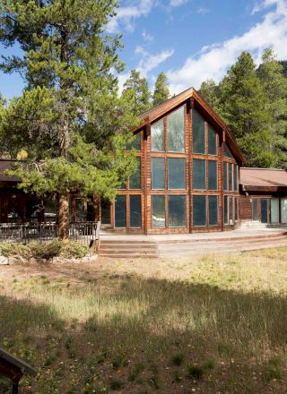 A rustic cabin-style lodge with large glass windows, wooden siding, and a spacious grassy yard surrounded by trees and mountains, under a bright blue sky.