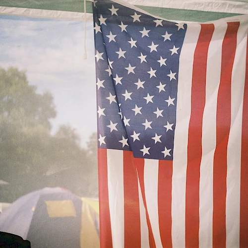 A tall American flag hangs indoors with trees and tents visible in the background, suggesting a campsite or outdoor event.