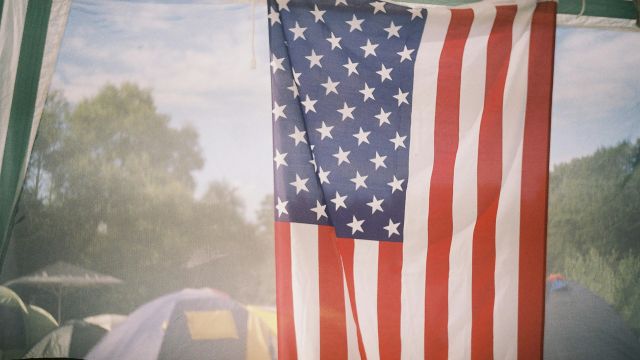 A tall American flag hangs indoors with trees and tents visible in the background, suggesting a campsite or outdoor event.