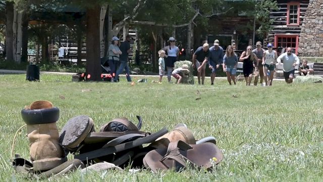 A pile of old hats and boots on grass with people walking and a wooden building in the background.