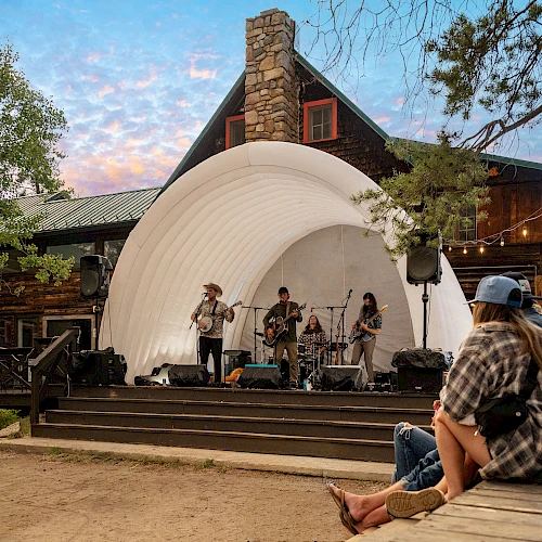 A small outdoor concert on a white shell stage at sunset, with a band playing as a casual audience sits on a wooden platform nearby.