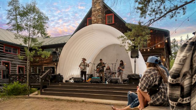 A small outdoor concert on a white shell stage at sunset, with a band playing as a casual audience sits on a wooden platform nearby.