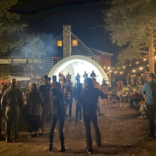 A nighttime outdoor gathering around a lit stage with people dancing and socializing near a white dome, string lights, and trees.