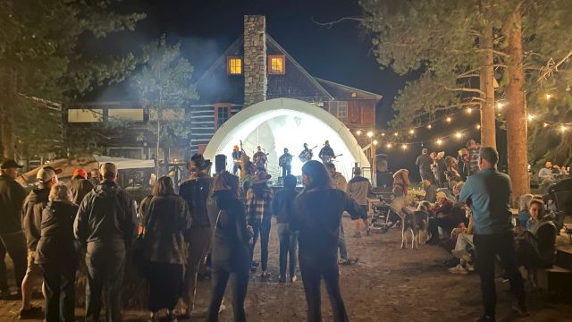 A nighttime outdoor gathering around a lit stage with people dancing and socializing near a white dome, string lights, and trees.