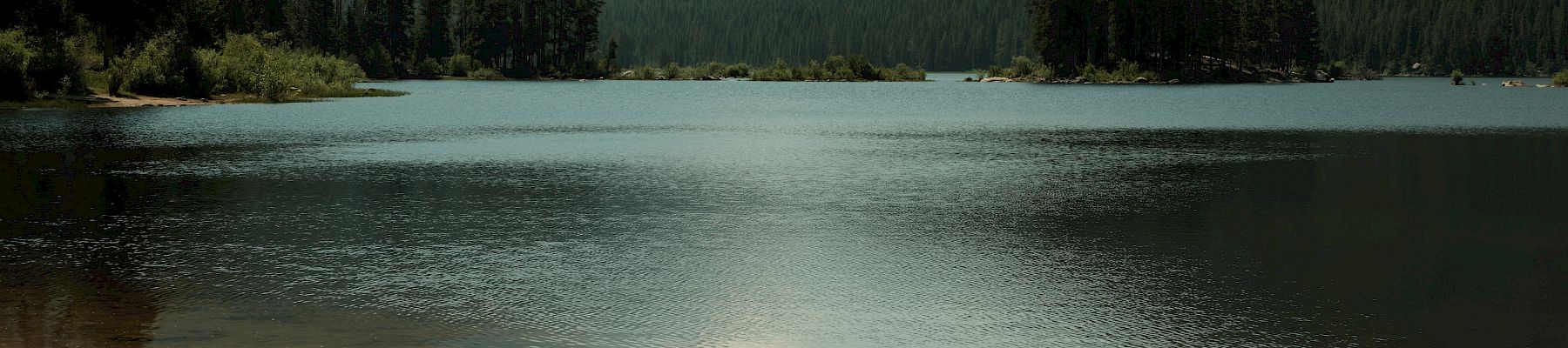 A calm mountain lake with pine forests and hazy peaks, plus a small inflatable kayak resting on the sandy shore.