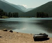 A calm mountain lake with pine forests and hazy peaks, plus a small inflatable kayak resting on the sandy shore.