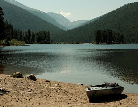 A calm mountain lake with pine forests and hazy peaks, plus a small inflatable kayak resting on the sandy shore.