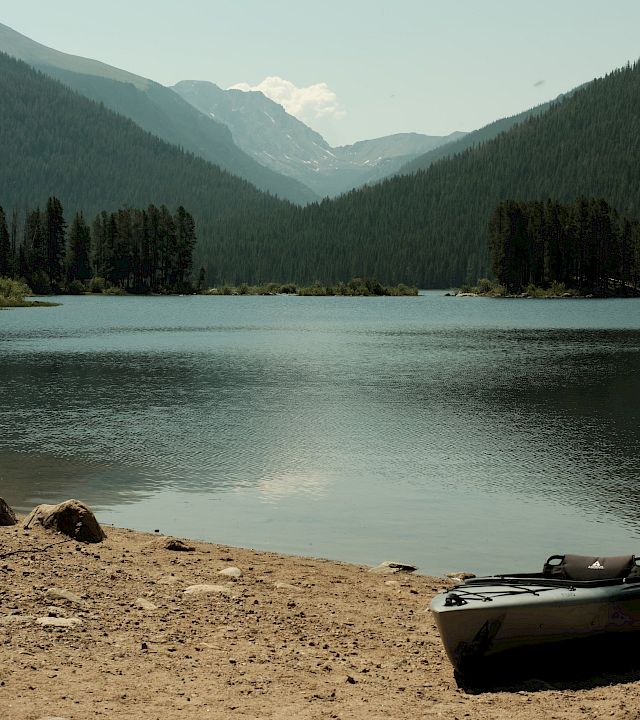 A calm mountain lake with pine forests and hazy peaks, plus a small inflatable kayak resting on the sandy shore.