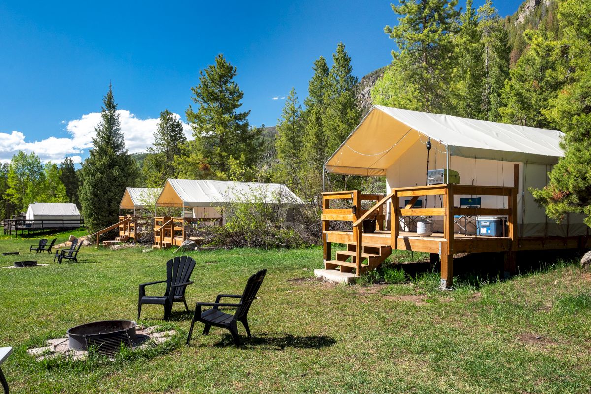 A sunny campground with wooden cabins and tents, pine trees, a fire pit, and plastic chairs around a grassy clearing, perfect for outdoors.
