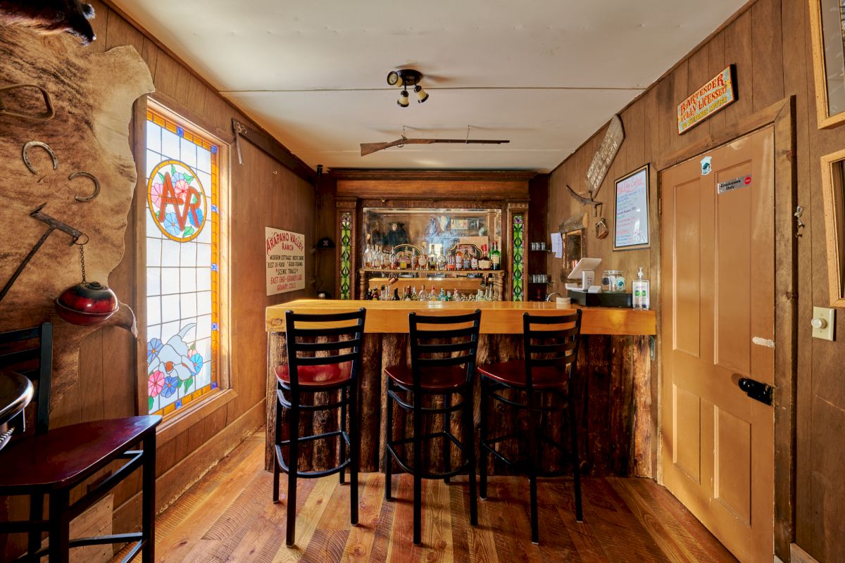 A cozy rustic bar with wooden floors, a long counter, stools, a backlit beer selection, chalkboards, and warm ambient lighting, inviting guests tonight.