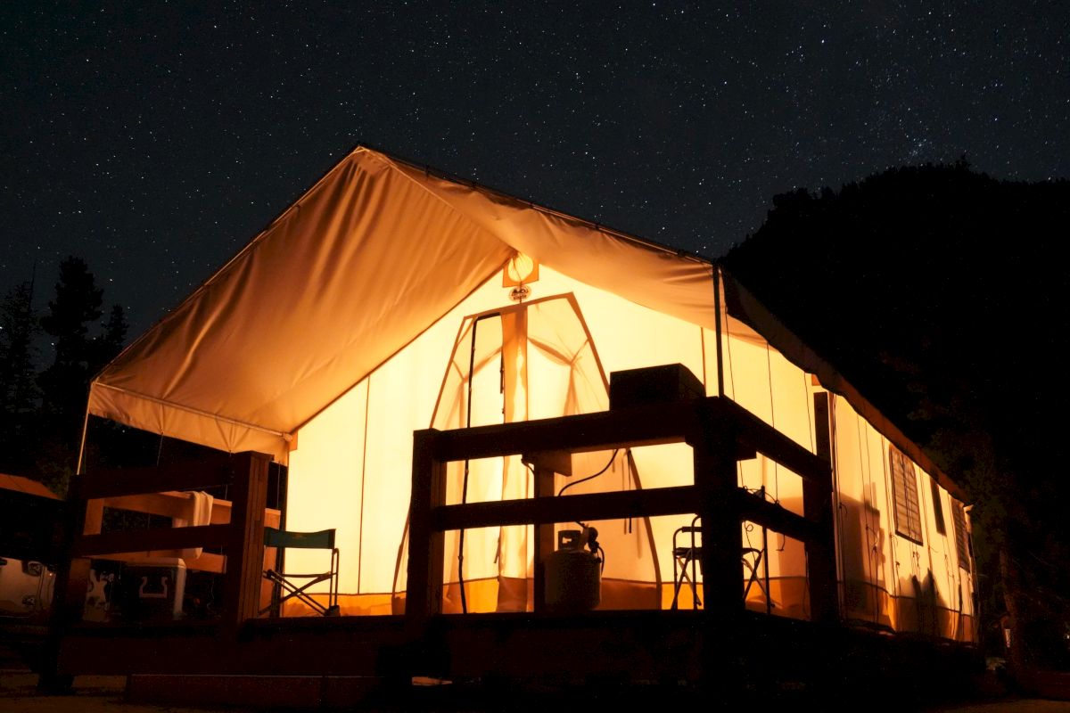 A glowing campsite tent at night with wooden furniture outside, a starry sky, and warm light illuminating the interior and deck.