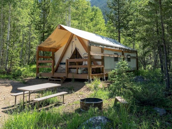 A wooden tent cabin in a forest clearing, with a small picnic table, a fire pit, and grassy surroundings under leafy trees.