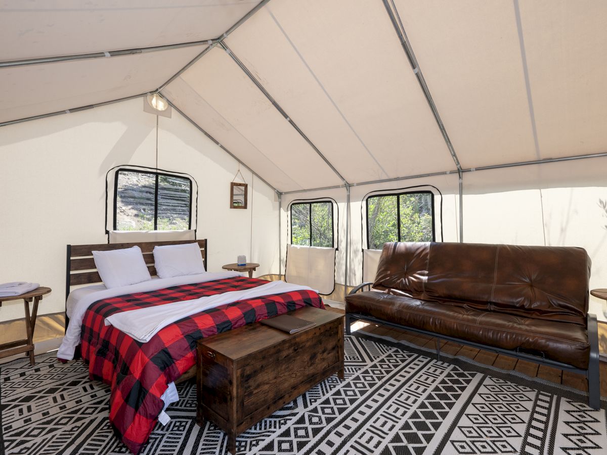 Inside a safari-style tent: a bed with red plaid blanket, two stools, and a brown leather sofa under skylight rails.