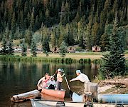 A group of people on a small orange boat near a lake, with others in blue boats, a wooden dock, and a forested hillside in the background, at a sunny day.